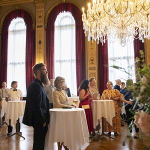 Ihmisiä suuressa salissa seisomassa pienten pyöreiden pöytien ääressä.