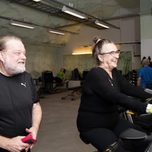 nainen polkee kuntopyörällä, mies katsoo vieressä.