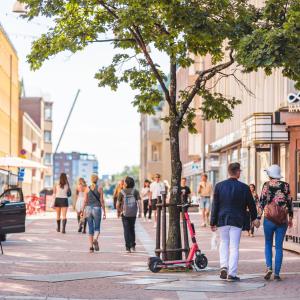 Ihmisiä kävelee kesäaikaan kadulla. Etualalla puu, johon nojaa sähköpotkulauta.