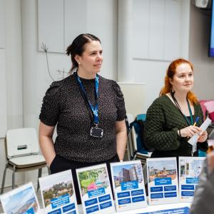 Two experts from the City of Tampere discuss with people at a residents&#039; evening. There are several display stands on the table.