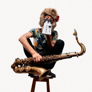 Tomoki Sanders seated on a stool holding a saxophone, wearing a floral shirt and a furry hat with flower. Their face is obscured by a small electronic device and an animal-themed mask.