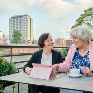 Kaksi ikääntynyttä henkilöä istuu pöydän ääressä edessään tablet-laite ja kahvikuppi.