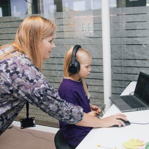 A little blonde-haired girl is sitting at a computer with headphones in her ears. Behind her is her mother, holding a computer mouse on the desk.
