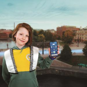 Poika seisoo Tampere-maiseman edessä ja näyttää kameralle Tampere.Finland -sovellusta.