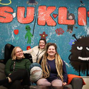 Kolme henkilöä istuu sohvalla ja hymyilee. Takana on seinä, jossa on Haisulin kuva ja jossa lukee Suklis.