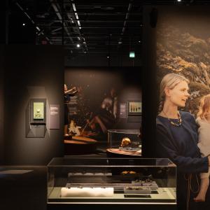 Hämärässä näyttelytilassa esineitä vitriinissä keskellä huonetta. Seinällä kuvassa äiti ja lapsi.