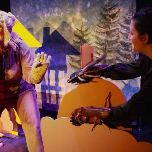 Koiran roolipuvussa oleva ihminen pitää toista &quot;tassua&quot; suun edessä ja hymyilee. Taustalla kuusia sekä varjokuva talosta.