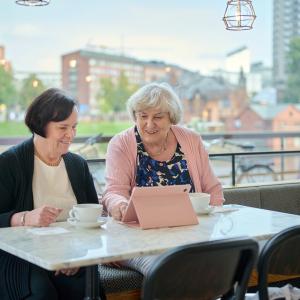 Two people are sitting in a café, smiling as they look at a tablet device.