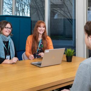 Kuvassa kolme henkilöä. Kaksi istuu pöydän toisella puolella kasvot kuvaajaan päin ja yksi selin. Kaikki hymyilevät. Heidän välissään on tietokone ja menossa on jonkinlainen ohjaustilanne.