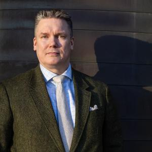 Henkilö seisoo tummassa puvussa tumman seinän edessä. Vieressä näkyy tumma varjo.