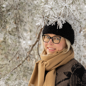 Henkilö hymyilee talvivaatteissaan lumisen puun alla.