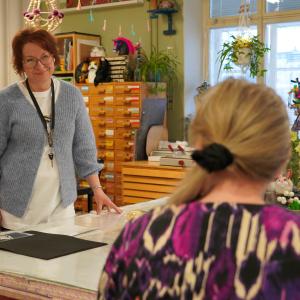 Henkilö seisoo pöydän ääressä. Etualalla toinen henkilö, joka tekee käsitöitä.