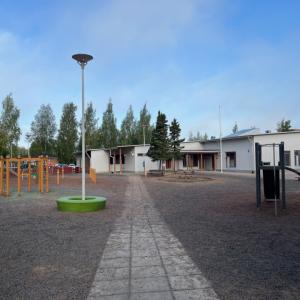Metsäniityn päiväkodin ja koulun pihaa, taustalla näkyy rakennus. Piha on lähinnä avointa sorakenttää, jossa on puisia leikkivälineitä. on 