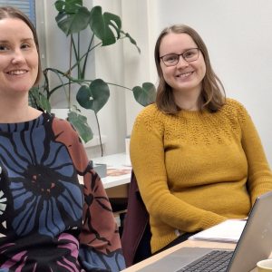 Kuvassa kaksi naista istumassa pöydän ääressä. Molemmat hymyilevät.