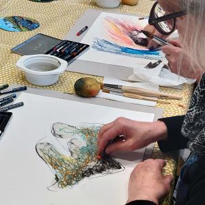 Harmaahiuksinen nainen maalaa värikkäillä liiduilla abstraktia teosta paperille.