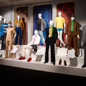 Miesten eri aikakauden vaatteisiin puetut mallinuket korokkeella näyttelytilassa.  