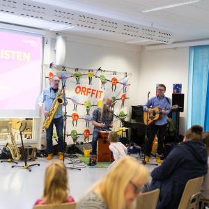 Lastenmusiikkiyhtye soittaa koulun tiloissa. Yleisössä on eri-ikäisiä ihmisiä.