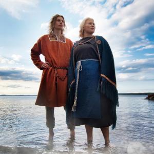 Mies ja nainen seisovat avojaloin matalassa vedessä, josta heijastuu sininen taivas. Pari on pukeutunut muinasipukuihin, naisella on sininen asu ja miehellä oranssi..