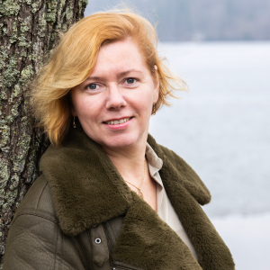 Henkilö nojaa puunrunkoon ja katsoo suoraan kameraan.