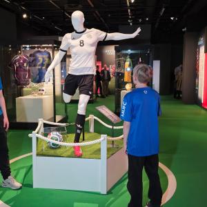 Kaksi lasta katselee mallinukkea, jonka päällä on jalkapalloasu näyttelytilassa.