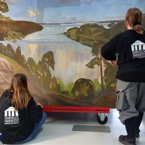 Kaksi mustapuseroista henkilöä tarkastelee isoa maisemamaalausta. 