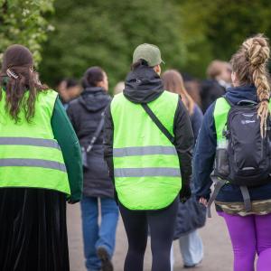 Kolme henkilöä kävelee rinnakkain vihreät huomioliivit yllään ihmisten keskellä.