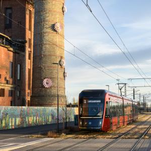 Kuvassa keskellä ratikka, vasemmalla piippu jossa kukiksta muodostuva teos.