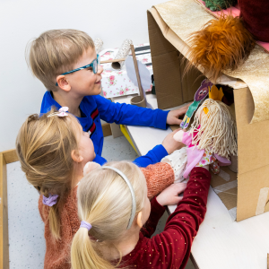 Three children play puppet theatre holding two puppets inside a cardboard box.