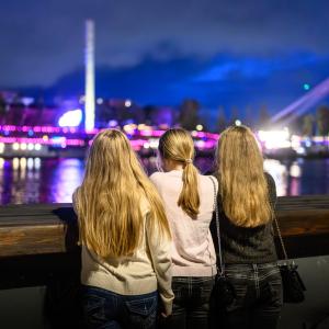Kolme nuorta nojaa kävelysiltaan selät kameraan päin iltaisessa Tampereessa.