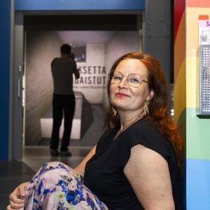 A red-haired woman in a black short-sleeved blouse is leaning against the wall. In the background, a museum display case stands out.