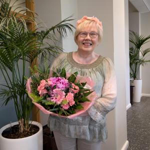 Aija Karttunen, with her light hair and a smile on her face, is standing next to a potted plant. She is holding a large bouquet of light pink flowers.