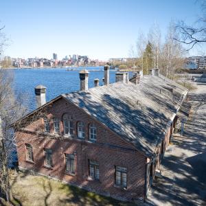 Ilmakuvan etualalla kuva punatiilisestä pesularakennuksesta. Taustalla näkyy Pyhäjärveä ja keskustan siluetti.