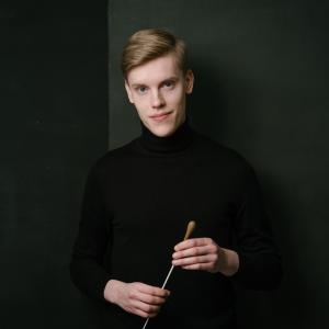 Conductor Kristian Sallinen in a black polo shirt, baton in hand, against a black background.