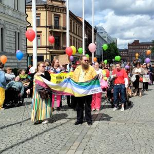 A procession of disabled people walks through Keskustori Central Square. Those in front hold a pride flag in their hands. Many are holding colourful balloons. Among the participants are wheelchair users.