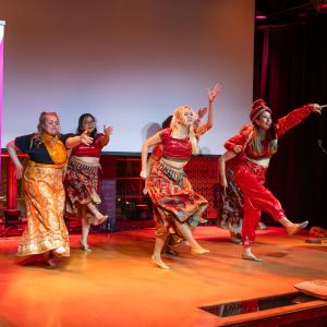 A group of dancers dressed in traditional ethnic costumes is performing on stage. At the edge of the stage is a roll-up banner for "Tampere Residents' Evenings."