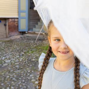 Pikkutyttö jurkistaa valkoisen lakanan takaa. Taustalla Amurin museokorttelin taloja.