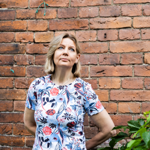 A person wearing a floral shirt is standing in front of a brick wall. Next to them are leaves from a bush.