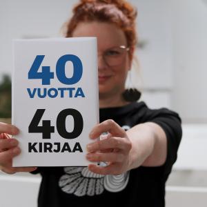 Tuija seisoo Metson parvikerroksessa ja pitää osittain kasvojensa edessä kirjaa, jonka kannessa on teksti 40 vuotta, 40 kirjaa. 