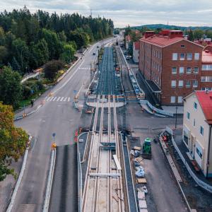 Kuvassa keskellä raitiotietyömaa ja autokaistoja, vihreää vasemmalla ja punatiilinen koulu oikealla. 