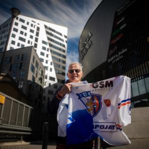 Jääkiekkovalmentaja Rauno Korpi näyttää pelipaitaa. Hänen takanaan näkyy Nokia Arenan lisäksi kerrostalo.