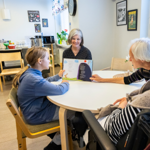 Lapsi näyttää vanhukselle kirjasta lukemaansa sivua. Opettaja ja toinen vanhus seuraavat.