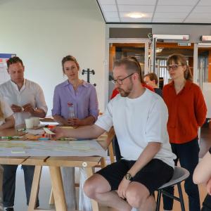 Henkilöt osallistuvat ryhmätyöhön ja ovat kerääntyneet pöydän ympärille. Pöydällä on muistiinpanoja ja kirjoitusvälineitä.