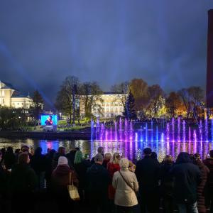 Yleisö seuraa iltahämärässä Tammerkosken rannalla Tanssivia vesiä. Veden pinnasta nousevat sinisin ja violetin sävyin valaistut suihkulähteet, taustalla näkyy valaistuja rakennuksia, tehdaspiippu sekä taivaalle suuntautuvia valokeiloja.