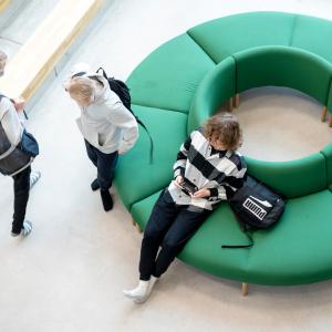 A top-down view of a round green sofa with one student sitting on it. Two other students are standing nearby.