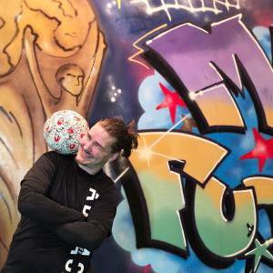 Mies temppuilee jalkapallon kanssa. Taustalla värikäs graffiti maalaus, jossa teksti MM Futis.