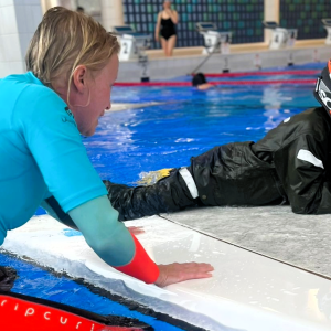 A woman leans over the edge of a pool while a child in a helmet lies on a floating device.