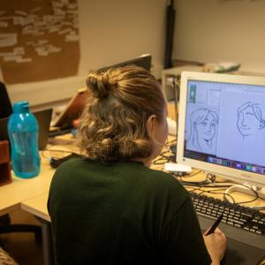 Kaksi henkilöä istuu työpöytien äärellä. Toisen tietokoneen ruutu näkyy kameralle, ja ruudulla on piirroksia.