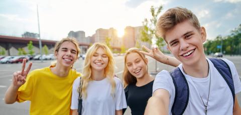 Neljä nuorta, kaksi naista ja kaksi miestä, seisovat parkkipaikan laidalla. Yksi ottaa selfie-kuvan, muut näyttävät käsimerkkejä ja hymyilevät. On kesä.