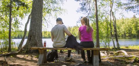 Nuori poika ja tyttö istuvat penkillä järven rannalla, heidän välissään koira.