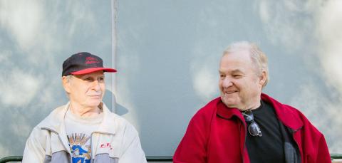 Kaksi miestä istuu ulkona seinän viereen asetetulla penkillä.